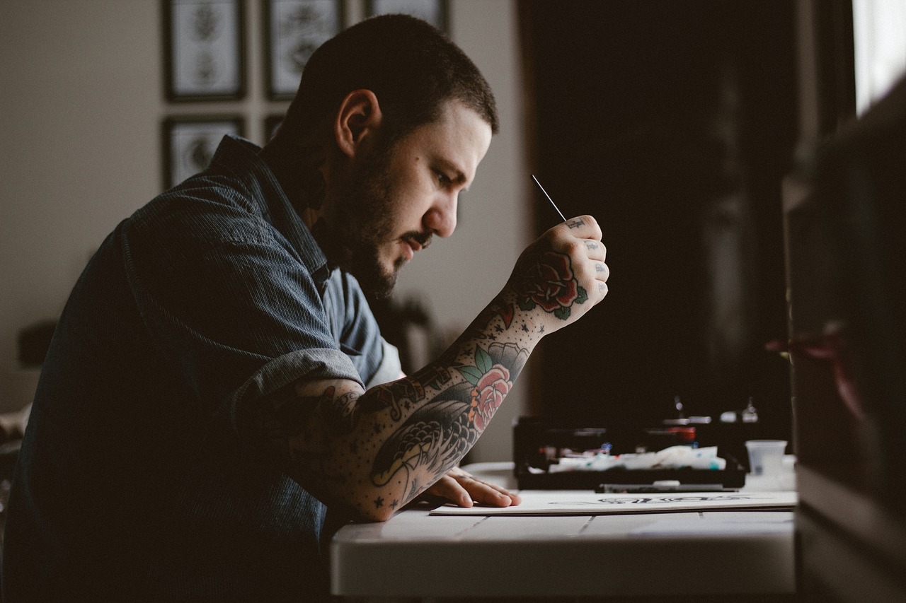 A tattooed artist bent over a drawing desk in window light