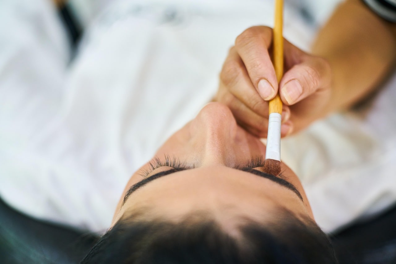 An artist working over a client on the treatment bed, brush in hand at the brow